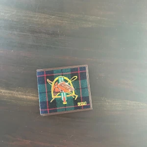 Rutig plånbok från Polo Ralph Lauren - Säljer en stilren plånbok från Polo Ralph Lauren med ett klassiskt rutigt mönster i grönt, blått och rött. Plånboken har flera fack för kort och är tillverkad i skinn med en elegant design. Perfekt för att hålla ordning på dina kort och kontanter.
