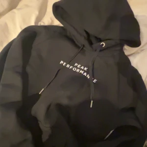 Svart hoodie från Peak Performance - Säljer en stilren svart hoodie från Peak Performance med vit logga på bröstet. Den har en klassisk design med dragsko i huvan och en bekväm passform. Perfekt för en avslappnad look. Köpte den på ztadium för inte så länge sedan men har aldrig kommit till användning (lappen är inte kvar) har testat en gång inget mer, köpte för 1000 något därav lite högre pris i och med att den också är typ helt ny, väldigt bra kvalité!