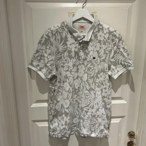 Vit blommig pikétröja från Levi's - Säljer en snygg vit pikétröja från Levi's med ett stilrent blommönster. Tröjan har korta ärmar och en klassisk krage med knappar. Perfekt för en avslappnad stil.