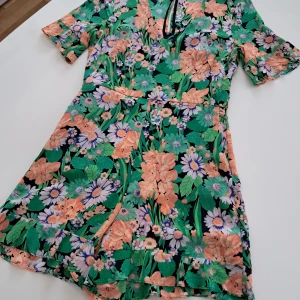 Blommig byxdress från Sandro Paris (Siden) - Snygg blommig kortklänning från Sandro Paris med färgglada blommor i grönt, rosa och blått. Byxdressen har korta ärmar och en v-ringad design med knappar framtill. Perfekt för en somrig look! Material, - SIDEN av högsta kvalitet. 