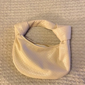 Beige axelväska med flätat mönster - Säljer en beige väska med snyggt flätat mönster och twistad axelrem. Väskan har dragkedja upptill och är gjord i ett mjukt syntetmaterial. Perfekt storlek för det viktigaste och ger en trendig touch till din outfit.