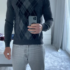 Mörkgrå argyle stickad tröja M - Snygg mörkgrå stickad tröja med klassiskt argylemönster i svart och grått framtill. Tröjan har rund halsringning och långa ärmar, perfekt för lager-på-lager. Passar dig som gillar stilren och tidlös look.