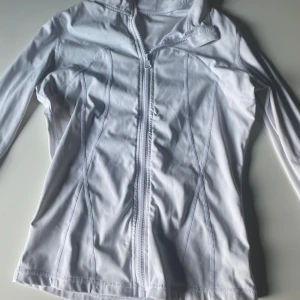 Vit sportig vindjacka med dragkedja - Snygg vit vindjacka med hel dragkedja framtill och figurnära passform. Jackan har långa ärmar och är tillverkad i ett lätt och smidigt material, perfekt för aktiva dagar. 