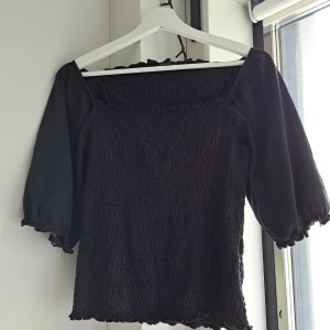 Svart blus med puffärm M 38 - Supersnygg svart topp i bomull från Medicine med smockad kropp och puffiga trekvartsärmar. Fyrkantig ringning och volangkant upptill och nedtill ger en trendig vibe. Perfekt att styla med jeans eller kjol för en cool look. Sparsamt använd.