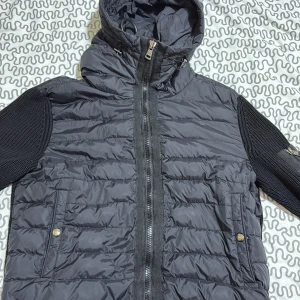 Svart pufferjacka från Moncler - Säljer en svart pufferjacka från Moncler med stickade ärmar och huva. Jackan har dragkedja framtill, två fickor med tryckknappar och Moncler-logga på ärmen. Kombinerar glansigt material på kroppen med grovstickade detaljer på ärmar och rygg för en riktigt clean look.
