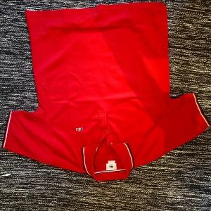 Röd pikétröja från Lacoste - Vad hände med kameran?  Snygg röd pikétröja från Lacoste med klassisk krage och knäppning framtill. Tröjan har broderad logga på bröstet och vita samt marinblå ränder på kragen och ärmsluten. Perfekt för en sportig och stilren look. Storlek L