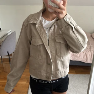 Beige jeansjacka från Perfect Jeans - Säljer en beige jeansjacka från Perfect Jeans i storlek S. Jackan är croppad med klassisk krage, två stora bröstfickor med lock och knappar framtill. Perfekt för dig som gillar en clean och stilren look. Passformen är normal och materialet är tåligt denim.