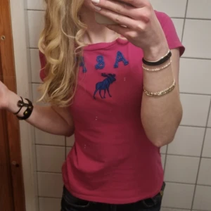 Rosa t-shirt från Abercrombie & Fitch - Snygg rosa t-shirt från Abercrombie & Fitch med blå broderad älg och bokstäver på bröstet. Klassisk passform och korta ärmar, perfekt till jeans eller shorts. Skön bomullskvalitet och stilren look för dig som gillar casual vibes.