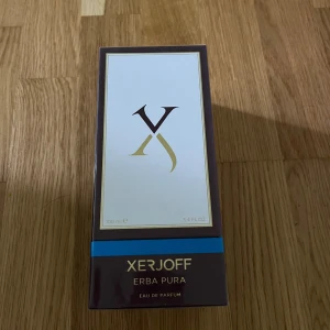 Xerjoff Erba Pura Eau de Parfum 100ml - Lyxig parfym från Xerjoff, Erba Pura, i en elegant mörklila och vit kartong med gulddetaljer och blå rand. Eau de Parfum, 100 ml, tillverkad i Italien. Förpackningen har en stilren och exklusiv design som verkligen sticker ut.