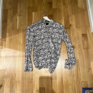 Blommig skjorta från Grand Frank - Snygg skjorta från Grand Frank med blommigt mönster i svart, vit, rosa och beige. Skjortan har lång ärm, klassisk krage och knäppning framtill. Perfekt för dig som vill sticka ut med en cool och färgglad look.