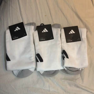 Adidas Milano 23 vita fotbollsstrumpor - Säljer Adidas Milano 23 fotbollsstrumpor i vitt med grå detaljer och svart Adidas-logga. Strumporna är knälånga och tillverkade i ett mjukt och ventilerande polyestermaterial med Aeroready-teknologi för att hålla fötterna torra under match eller träning.