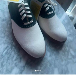 Polo Ralph Lauren Rhett Saddle skor - Snygga saddle shoes, som är special beställda  från Polo Ralph Lauren i vit mocka med svart detalj och klassisk snörning. Skorna har rund tå, platt sula i gummi och stilren kontrast mellan färgerna. Perfekta för dig som gillar preppy och tidlös stil.