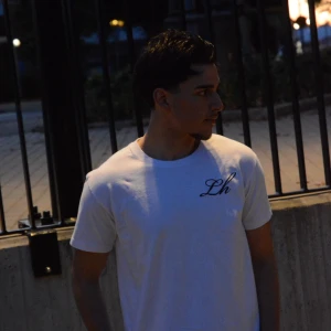 Vit t-shirt med tryck och Lh-logga - Vit t-shirt med svart Lh-logga på bröstet och stort grafiskt tryck på ryggen. Klassisk passform med rund hals och korta ärmar. Perfekt för dig som gillar stilrena plagg med en twist.