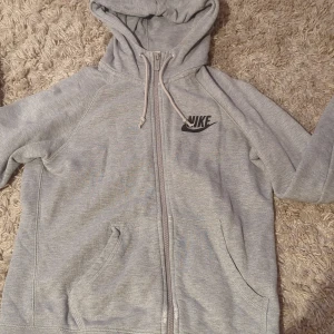Grå hoodie från Nike med dragkedja - Grå Nike hoodie med svart logga på bröstet, hel dragkedja och huva med snörning. Mjuk bomullsblandning och två fickor framtill. Perfekt för chill dagar eller när du vill ha en sportig look.