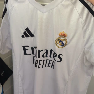 Real Madrid vit matchtröja Adidas - Snygg vit Real Madrid fotbollströja från Adidas med klubbmärke och svart Adidas-logga på bröstet. Tröjan har korta ärmar, svart tryck med Emirates sponsor och klassiska svarta detaljer på axlarna. Perfekt för dig som älskar fotboll och vill visa ditt stöd för laget. Rodrygo är på ryggen!