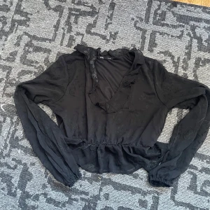 Svart blommig blus med volang - Supersöt svart blus från Sinsay med transparent tyg och diskret blommigt mönster. Blusen har v-ringning med volangkant, långa ärmar och resår i midjan för en snygg passform. Perfekt för dig som gillar romantiska detaljer och flowy vibes.