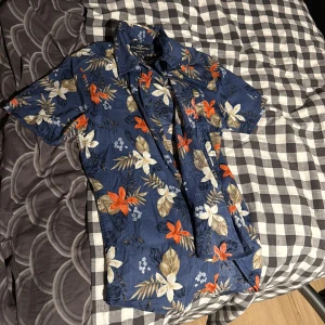 Blommig blå skjorta Hampton Republic - Snygg kortärmad skjorta från Hampton Republic i blått med stora blommor i vitt, orange och beige. Regular fit med klassisk krage och knappar framtill. Perfekt för sommarens alla äventyr och chill dagar.