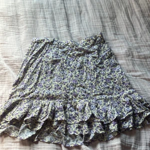 Blommig volangkjol från Lindex - Supersöt kort kjol från Lindex med blommigt mönster i lila och gult. Kjolen har volangkant nertill och resår i midjan för skön passform. Perfekt för vår och sommar när du vill ha en chill och färgglad look.