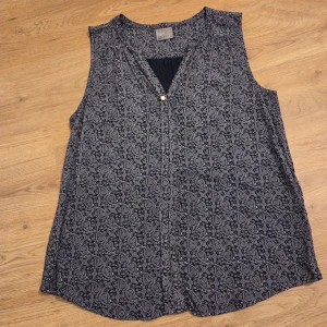 mönstrade önstrad ärmlös blus Vero Moda - Snygg ärmlös blus från Vero Moda i marinblå med vitt paisleymönster. Toppen har v-ringning med en liten knappdetalj framtill och är tillverkad i ett mjukt, lätt material som känns skönt mot huden. Perfekt för dig som gillar stilrena mönster och relaxed fit.