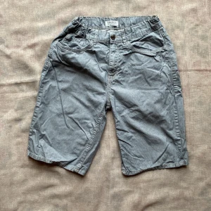 Randiga grå shorts från Lindex - Snygga grå shorts från Lindex med tunna vita ränder och klassisk femficksdesign. De har justerbar midja med knappresår och bälteshällor. Perfekta för sommardagar och har en avslappnad passform som ger skön känsla hela dagen.