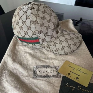 Gucci keps - Exklusiv keps från Gucci i beige canvas med klassiskt GG-monogram över hela ytan. Kepsen har justerbar rem i brunt läder och ikoniska gröna och röda ränder baktill. Perfekt accessoar för dig som vill sticka ut med lyxig streetstyle.😉