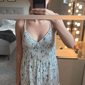 Vit blommig sommarklänning med smala band - Superfin vit kortklänning med blått blommönster och smala axelband som korsas i ryggen. Klänningen har v-ringning, markerad midja och ärmlös design. Perfekt för varma dagar och ger en fräsch och luftig känsla.