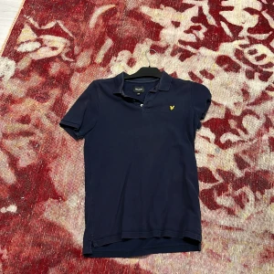 Marinblå pikétröja från Lyle & Scott - Snygg marinblå pikétröja från Lyle & Scott Junior med klassisk krage, korta ärmar och gul broderad logga på bröstet. Tröjan är i bomull och har knappar framtill för en clean look. Perfekt för dig som gillar stilren och sportig vibe.