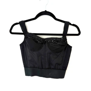 Svart korsettopp från Fox Isle of France - Snygg svart korsettopp från Fox Isle of France med breda axelband och spetsdetaljer upptill. Toppen har hakknäppning bak och är tillverkad i ett stretchigt syntetmaterial. Perfekt för att skapa en edgy look med extra attityd.