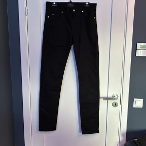 Svarta jeans från J.Lindeberg - Svarta jeans från J.Lindeberg med klassisk femficksdesign och silverfärgade nitar. Byxorna har en rak passform och är tillverkade i bomull med en liten mängd stretch för extra komfort. Snyggt diskret JL-logo på myntfickan.