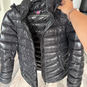 Svart dunjacka från Colmar, strl s/m - Snygg svart dunjacka från Colmar med glansig finish och klassisk pufferstil. Jackan har huva, dragkedja framtill och två fickor. Insidan är mönstrad med Colmar-loggor. Perfekt för kalla dagar och riktigt skön att bära.