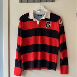 Randig långärmad pikétröja från Gant - Snygg långärmad pikétröja från Gant med röda och marinblå ränder, vit krage och broderad G på bröstet. Storleken på tröjan är 158/164 cm. Om du har några frågor skriv gärna. 