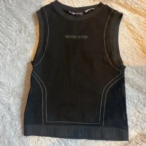 Svart träningslinne från FBSister XS - Cool svart ärmlös topp från FBSister med texten 'NEVER STOP' framtill. Toppen har ribbade kanter med tunna ränder och meshdetaljer på sidorna som ger en sportig vibe. Perfekt för dig som gillar en edgy och aktiv stil.