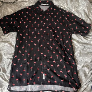 Svart skjorta med flamingomönster - Svart skjorta från Lager 157 med korta ärmar och helknäppning framtill. Skjortan har ett coolt mönster med rosa flamingos över hela tyget. Perfekt för dig som vill sticka ut med en lekfull stil. Säljer mycket skjortor så köp gärna flera i ett bundle!