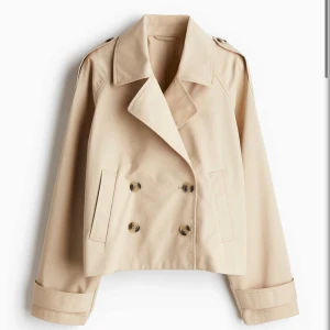 Beige croppad trenchcoat med knappar - Stilren croppad trenchcoat i beige med dubbelknäppning och bred krage. Jackan har långa ärmar, axelklaffar och raka fickor framtill. Perfekt för dig som vill ha en klassisk look med modern twist.