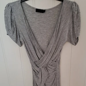 Grå omlottklänning från All Saints - Supersnygg grå kortklänning från All Saints med omlott och draperad effekt framtill. Puffiga kortärmar med diskreta pärldetaljer ger en unik touch. Mjuk och stretchig bomullskänsla, perfekt för dig som gillar stilrena men ändå speciella plagg.