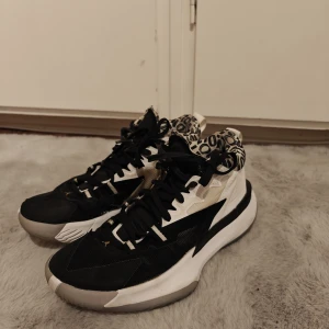 Svarta och vita Jordans basketskor - Snygga sneakers från Jordan i svart och vitt med coolt grafiskt mönster runt ankeln. Skorna har snörning, platt sula och detaljer i guld. Ovandelen är i syntet och textil, sulan är vit med grått mönster undertill. Basketskor, 7/10 skick. Skriv om ni har frågor!