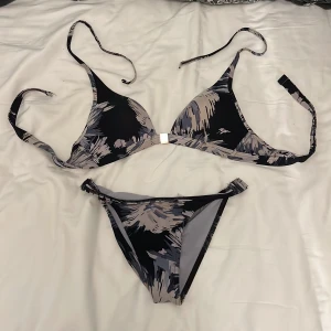Söt mönstrad bikini - Snygg bikini med svart bas och abstrakt mönster i beige och grått/blått. Överdelen har trekantskupor med knyt i nacken och rygg, samt en liten silverfärgad detalj framtill. Underdelen är klassisk med låg midja och matchande mönster. Perfekt för stranden eller poolen! OBS! Osäker på storleken då etiketten är bortklippt