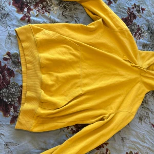 Gul hoodie med huva och ficka - Snygg gul hoodie med huva och stor magficka. Tröjan har långa ärmar och ribbade muddar vid ärmslut och nederkant. Perfekt för dig som gillar färg och vill sticka ut lite extra. Skön och mjuk bomullskänsla.