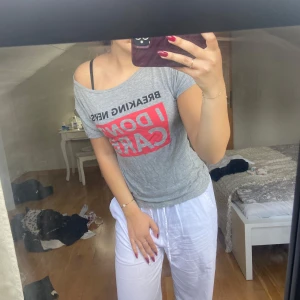 Grå off shoulder t-shirt med tryck "I DON'T CARE" - Snygg grå t-shirt med svart och rött tryck där det står "BREAKING NEWS: I DON'T CARE". T-shirten har korta ärmar och en avslappnad offshoulder-passform. Den är väldigt löss och skön.