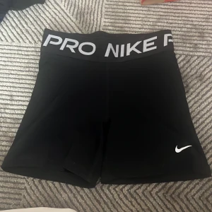 Nike Pro svarta träningstights-shorts - Svarta Nike Pro shorts med bred resår i midjan där det står 'NIKE PRO' i vitt. Tight passform och korta ben, perfekta för träning eller gym. Swoosh-logga i vitt på ena benet. Gjorda i stretchigt syntetmaterial som andas.