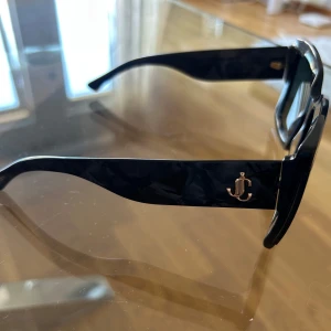 Jimmy chip sol glasögon  - Snygga svarta solglasögon från JC med breda skalmar och guldig JC-logga på sidan. Glasögonen har en klassisk, fyrkantig form och är tillverkade i blank plast. Perfekta för att lyfta din stil till nästa nivå.
