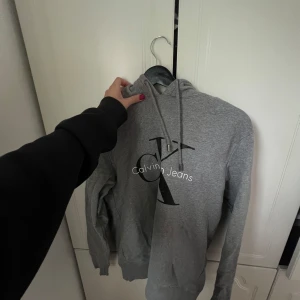 Grå Calvin Klein hoodie med logga - Snygg grå hoodie från Calvin Klein Jeans med stor svart och vit logga framtill. Klassisk modell med huva och snörning, ribbade muddar och mjukt material. Perfekt för dig som gillar stilrena och bekväma plagg med streetkänsla.