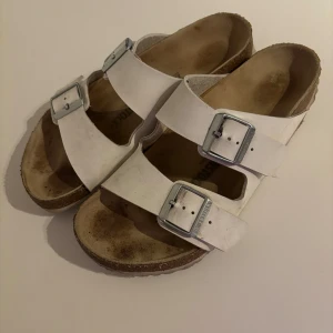 Vita sandaler från Birkenstock - Klassiska vita sandaler från Birkenstock med två breda remmar och stora silverspännen. Fotbädden är anatomiskt utformad och sulan är i kork för extra komfort. Perfekta för sommaren och riktigt sköna att gå i.