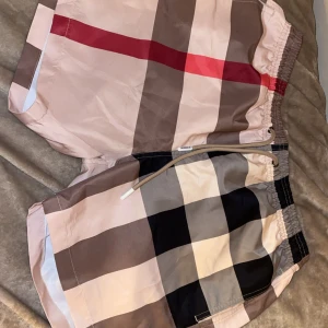 Rutig shorts med snörning Burberry - Snygga rutiga shorts från Burberry med klassiskt beige, svart, rött och vitt mönster. Shortsen har elastisk midja med snörning och är tillverkade i ett lätt och bekvämt material, perfekta för sommaren. Stilen är loose och de har en modern vibe. Finns i alla storlekar 