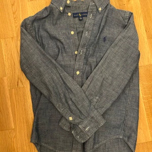 Blå skjorta från Ralph Lauren M - Klassisk blå skjorta från Ralph Lauren i bomull med button down-krage och broderad logga på bröstet. Skjortan har långa ärmar, vita knappar och en avslappnad passform. 