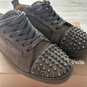 Bruna mockasneakers från Louboutin - Snygga bruna sneakers i mocka från Christian Louboutin med coola svarta nitar över tån. Skorna har svarta snören och en platt sula. Perfekta för dig som vill sticka ut med en edgy och lyxig stil.