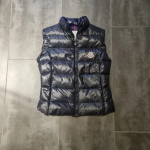 Mörkblå dunväst från Moncler - Snygg mörkblå dunväst från Moncler med glansig finish och klassisk logga på bröstet. Västen har hög krage, quiltad design och dragkedja framtill. Perfekt för lager-på-lager och ger en clean look till din outfit.