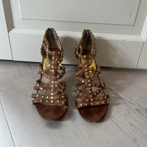 Bruna klackskor i sandalstil med nitar från Michael Kors Kors - Snygga bruna öppna klackskor från Michael Kors med smala remmar dekorerade med guldiga nitar. Skorna har öppen tå, smal klack och dragkedja bak. Tillverkade i skinn och perfekta för dig som vill sticka ut med detaljer.