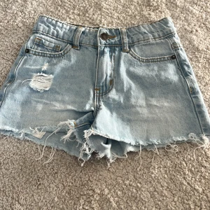 Ljusblå jeansshorts från UNIQ - Snygga ljusblå jeansshorts från UNIQ med fransig, rå kant och slitningar framtill. Klassisk femficksmodell med knapp och dragkedja. Perfekta för varma dagar och ger en avslappnad vibe. Hålet på andra bilden har jag it gjort själv är så i modellen. För barn