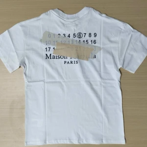 Vit Maison Margiela t-shirt med tryck - Vit t-shirt från Maison Margiela med svart tryck på både bröstet och ryggen. Klassisk rund hals och korta ärmar. T-shirten är i mjuk bomull och har den ikoniska siffergrafiken på ryggen samt diskret logga framtill. Perfekt för dig som gillar stilren design med en twist.